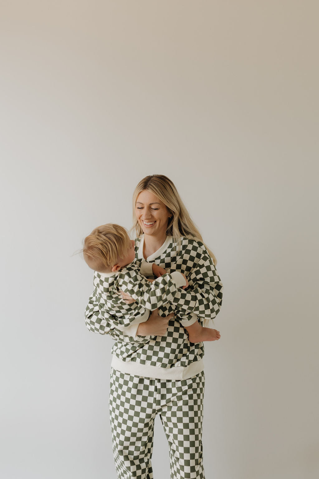 A woman beams as she holds a toddler, both in matching 'Adult Sweat Set | Lucky' by forever french baby, perfect for cooler weather. They pose against a light-toned backdrop.