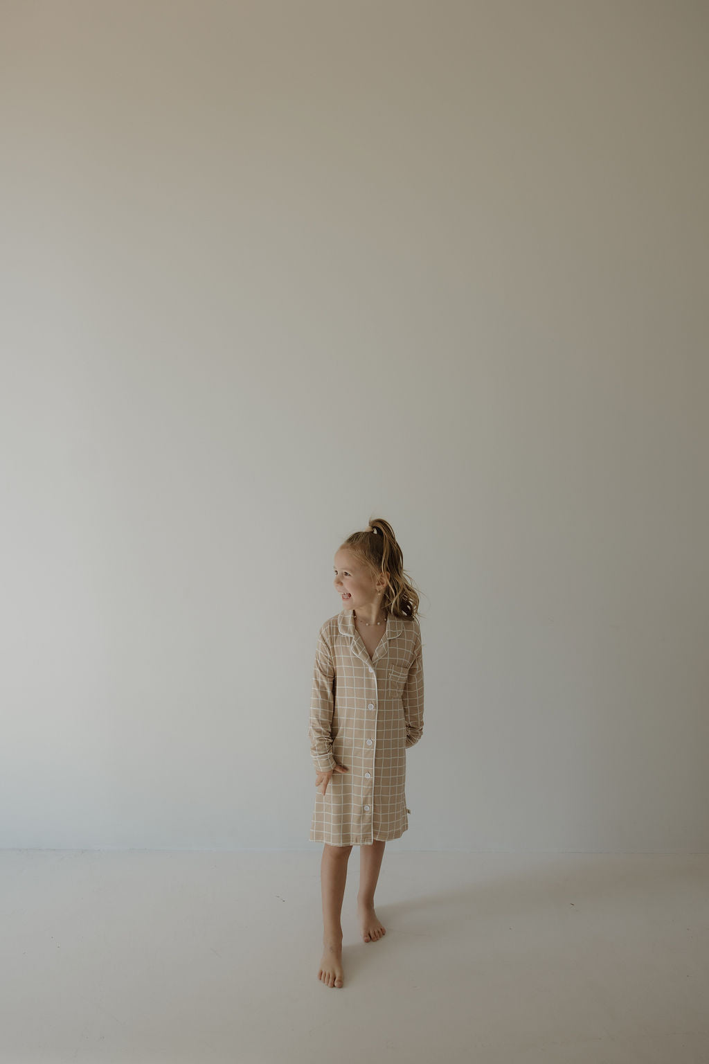 A young blonde girl stands barefoot on a light floor, wearing the forever french baby Toddler Bamboo Sleeping Dress | Grid, looking to her right against a plain, neutral wall.