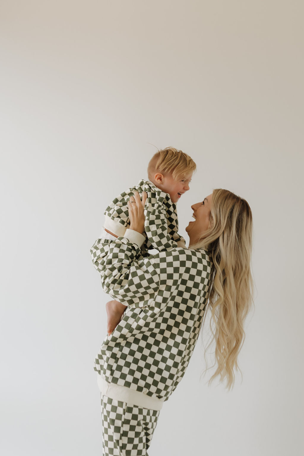 A woman with long blonde hair smiles holding a baby, both wearing matching black and white checkered rompers from the Adult Sweat Set | Lucky by forever french baby, standing against a plain, light-colored background that highlights their coordinated charm.