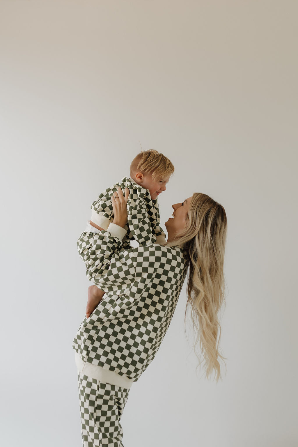 A woman with long blond hair holds up a toddler, both wearing matching forever french baby's black and white checkered "Lucky" sweat sets. They smile warmly at each other against a plain white background, perfect for cooler weather.