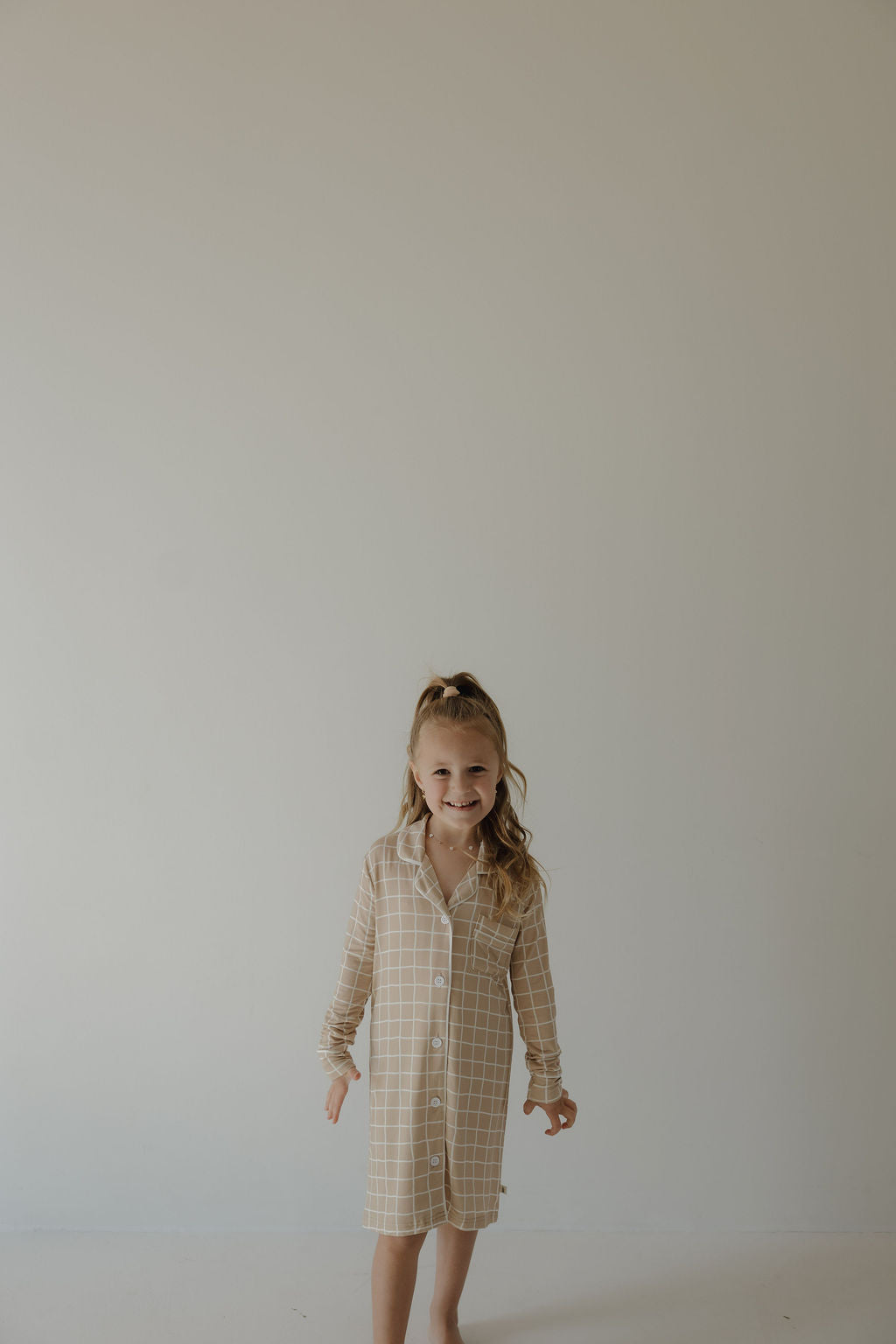 A young girl with long hair smiles against a plain, light wall, wearing the forever french baby Toddler Bamboo Sleeping Dress | Grid—crafted from soft, hypoallergenic bamboo fabric that's perfect for kids.