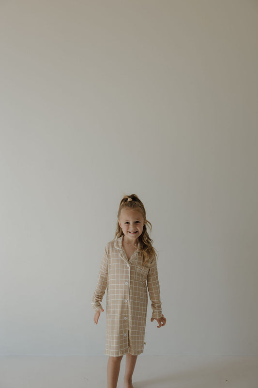 A young girl with long hair smiles against a plain, light wall, wearing the forever french baby Toddler Bamboo Sleeping Dress | Grid—crafted from soft, hypoallergenic bamboo fabric that's perfect for kids.