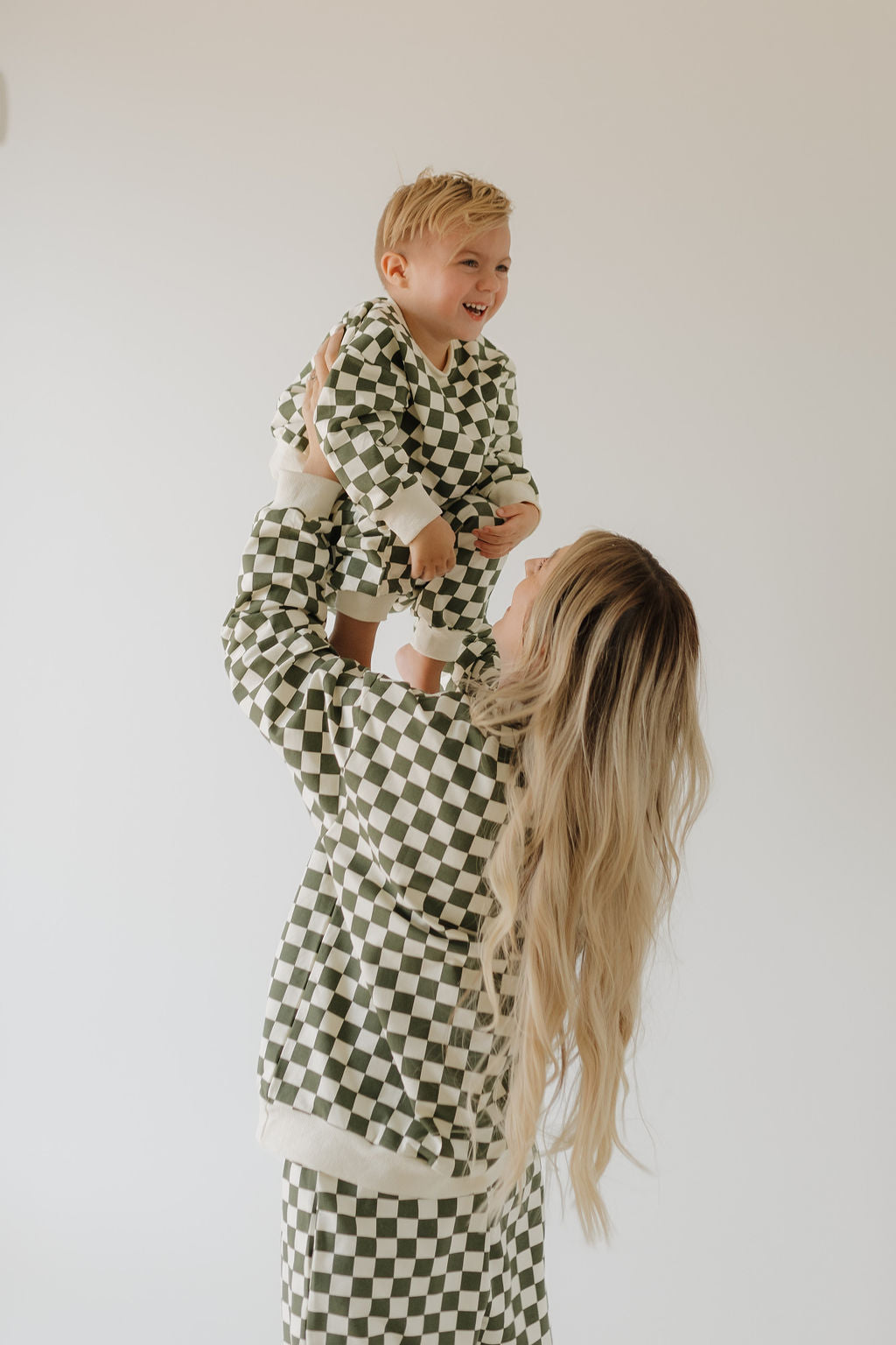 A woman with long blonde hair lifts a smiling child, both wearing the "Sweat Set | Lucky" from forever french baby in green and white checks. A plain, light-colored wall accentuates their joyful connection.