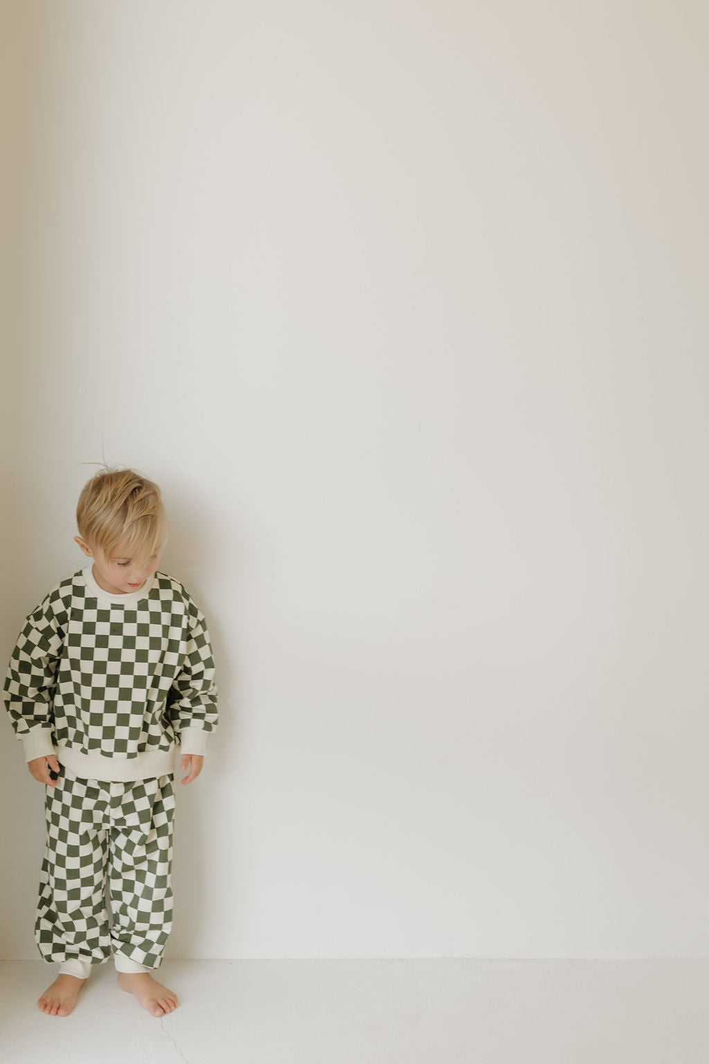 A young child stands against a plain wall, wearing a Sweat Set | Lucky by forever french baby. The child's gaze is lowered, hands relaxed at their sides. The room has a minimalist aesthetic, creating a calm and neutral background.