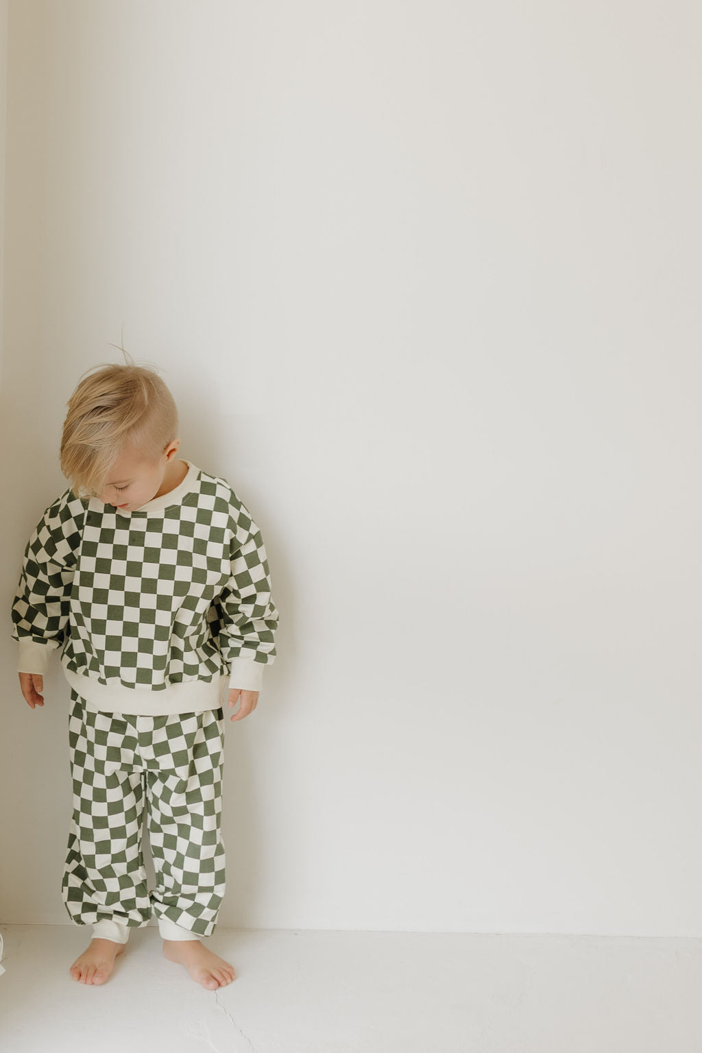 A young blond child stands barefoot against a white wall, dressed adorably in the "Sweat Set | Lucky" by forever french baby. The green and white checkered design mirrors charming adult matching sets, with the child gazing downward in a relaxed posture.