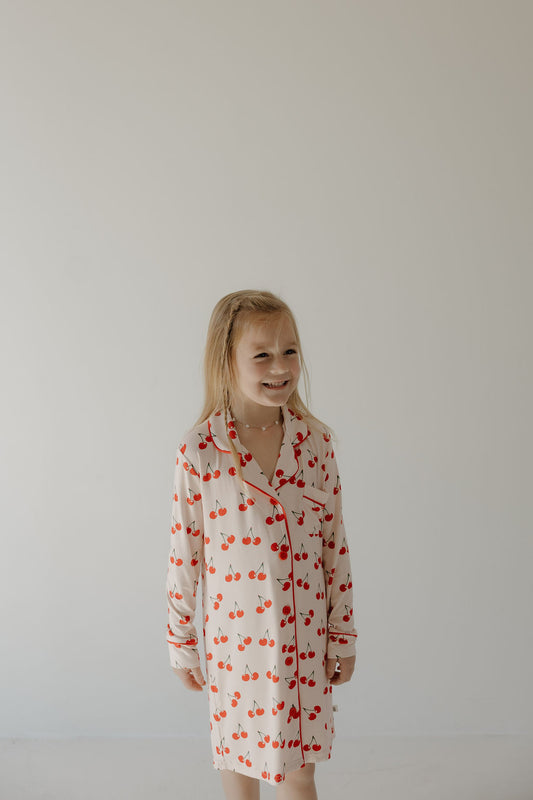 A young girl with long blonde hair smiles while wearing the forever french baby Toddler Bamboo Sleeping Dress | Cherry Orchard, featuring a cherry pattern, against a plain light background.