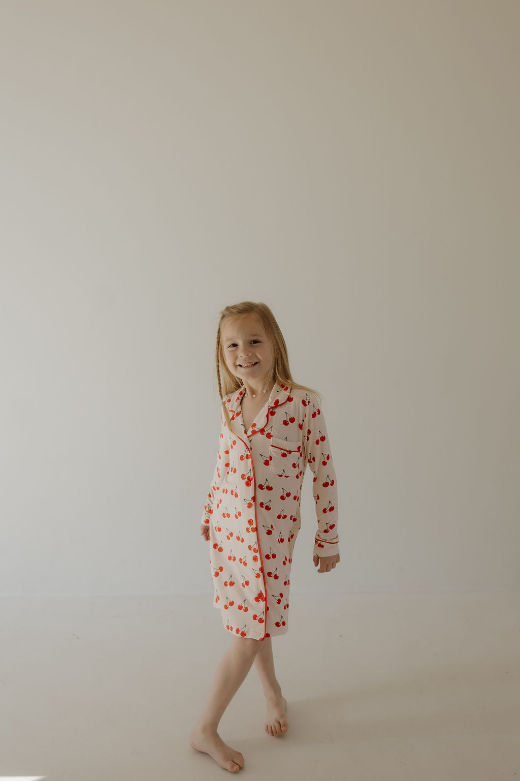 A young blond girl smiles barefoot in a bright room, wearing the forever french baby Toddler Bamboo Sleeping Dress | Cherry Orchard. This hypo-allergenic dress with a cherry pattern and red trim keeps her comfy all night.