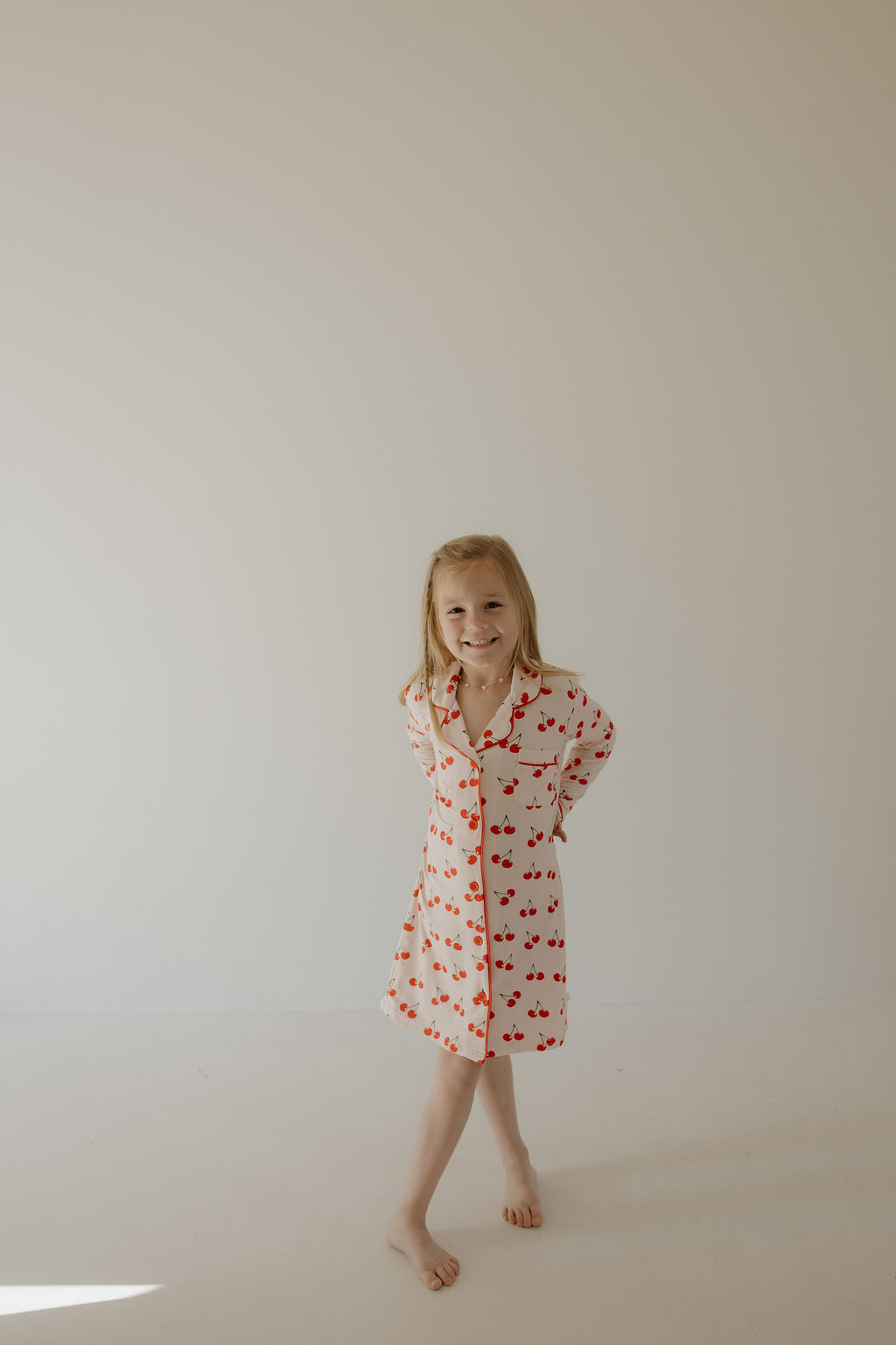 A young blonde girl smiles barefoot on a white floor against a light wall, wearing the forever french baby Toddler Bamboo Sleeping Dress | Cherry Orchard—hypoallergenic, comfortable, and charming with its sweet red heart pattern.