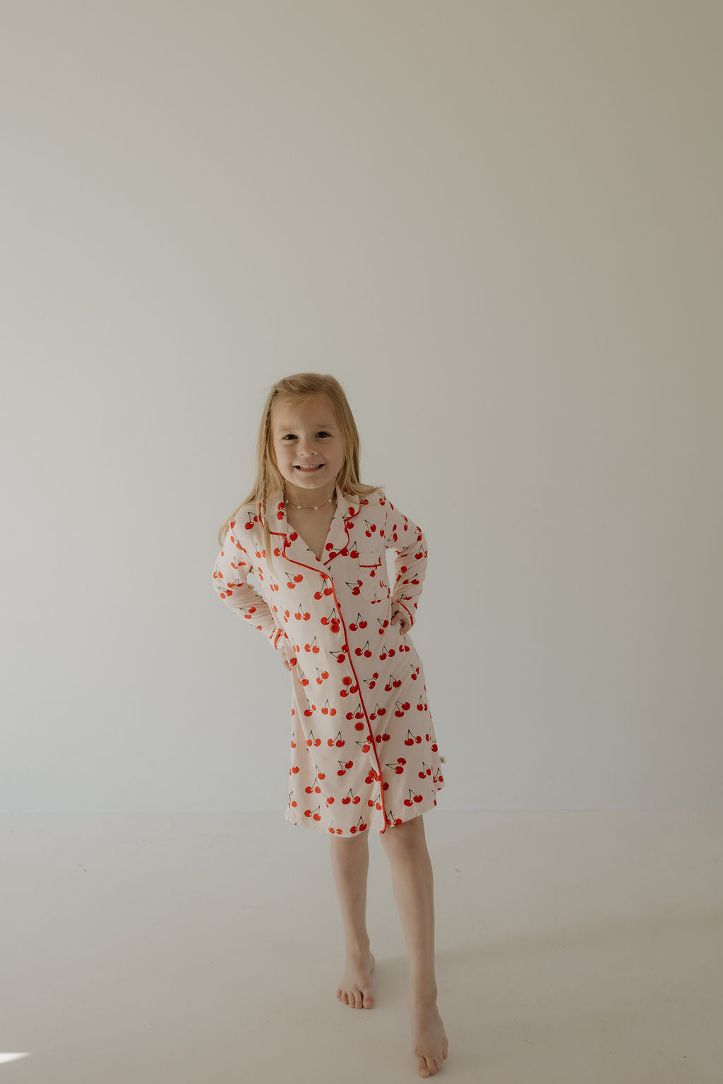A blonde toddler smiles barefoot on a white floor, wearing the forever french baby Toddler Bamboo Sleeping Dress | Cherry Orchard—soft, hypo-allergenic, and perfect for sensitive skin—set against a plain light background.