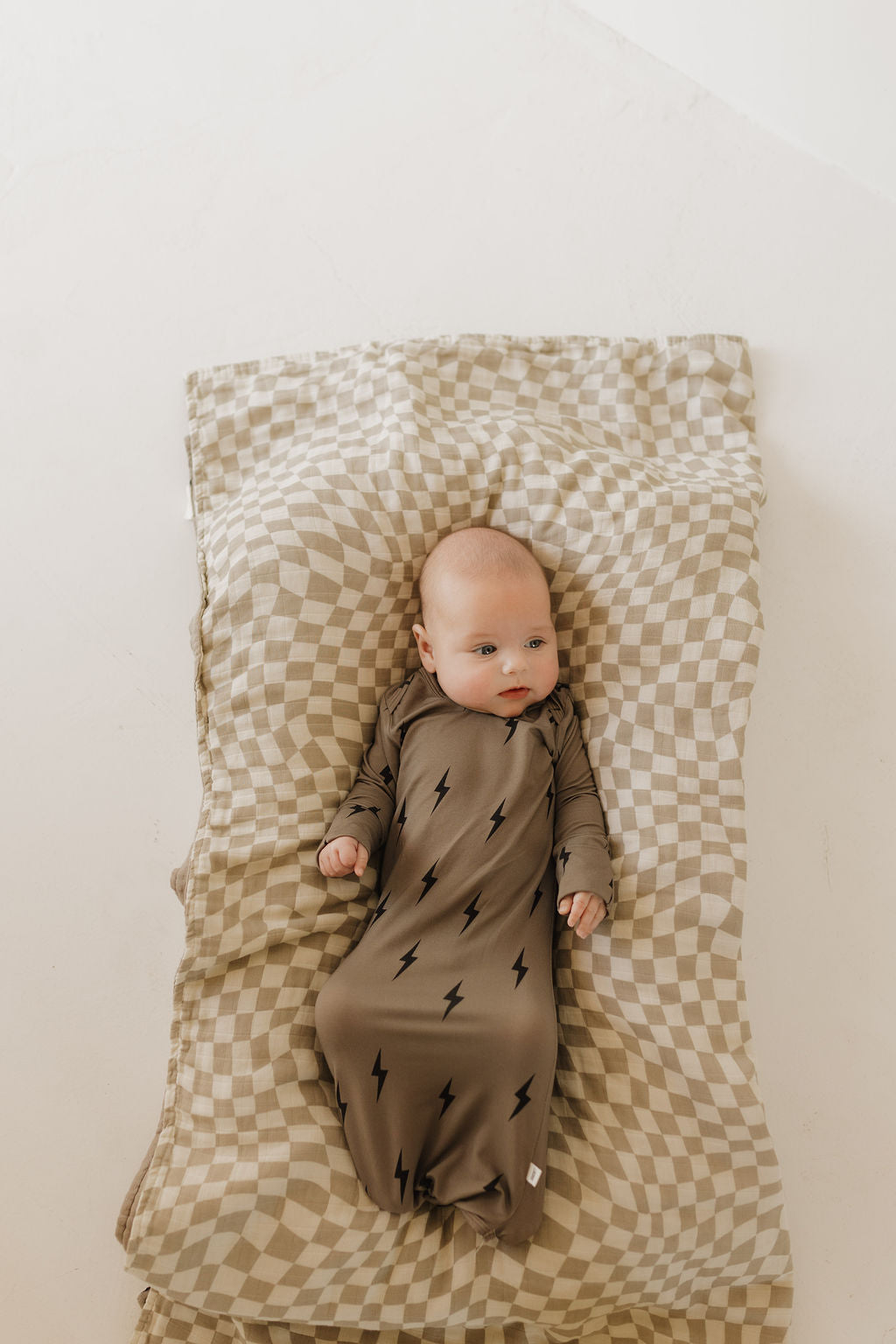 A baby, dressed in a forever french baby Bamboo Knotted Gown in brown with black lightning bolts, rests on a beige and white checkered blanket, the outfit gently contrasting with the light background as the baby gazes to the side.