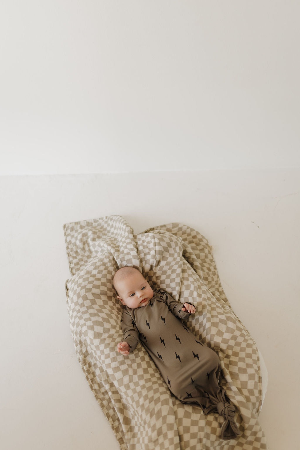 A baby in the forever french baby Bamboo Knotted Gown, featuring a brown and black lightning bolt pattern, lies on a beige and white checkered blanket. The breathable, hypo-allergenic fabric offers comfort against the plain, light-colored backdrop.