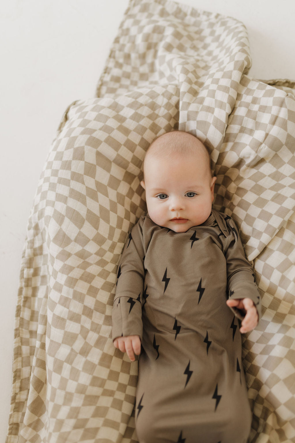 A baby lies on a beige and white checkered blanket, wearing the forever french baby Bamboo Knotted Gown in brown with black lightning bolt patterns. The breathable bamboo material ensures comfort as the baby gazes at the camera calmly.