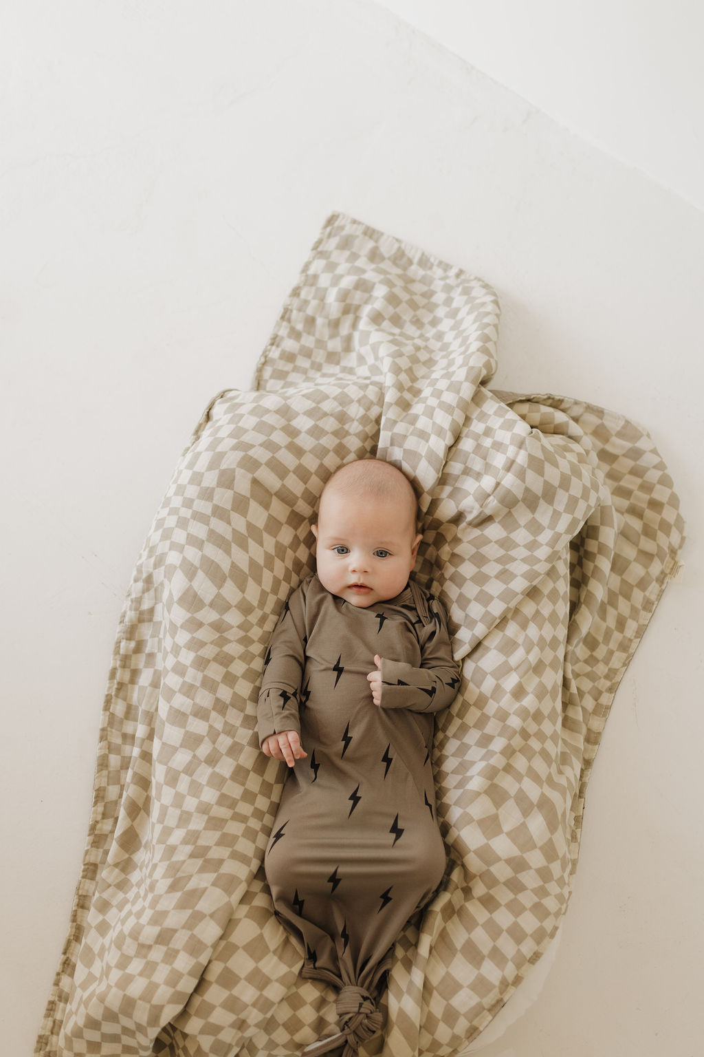 A baby, wrapped in breathable comfort, lies on a beige and white checkered blanket while wearing the "Bamboo Knotted Gown | Brown & Black Lightning Bolt" by forever french baby. The hypo-allergenic blanket enfolds the baby against the light background as they gaze upward.