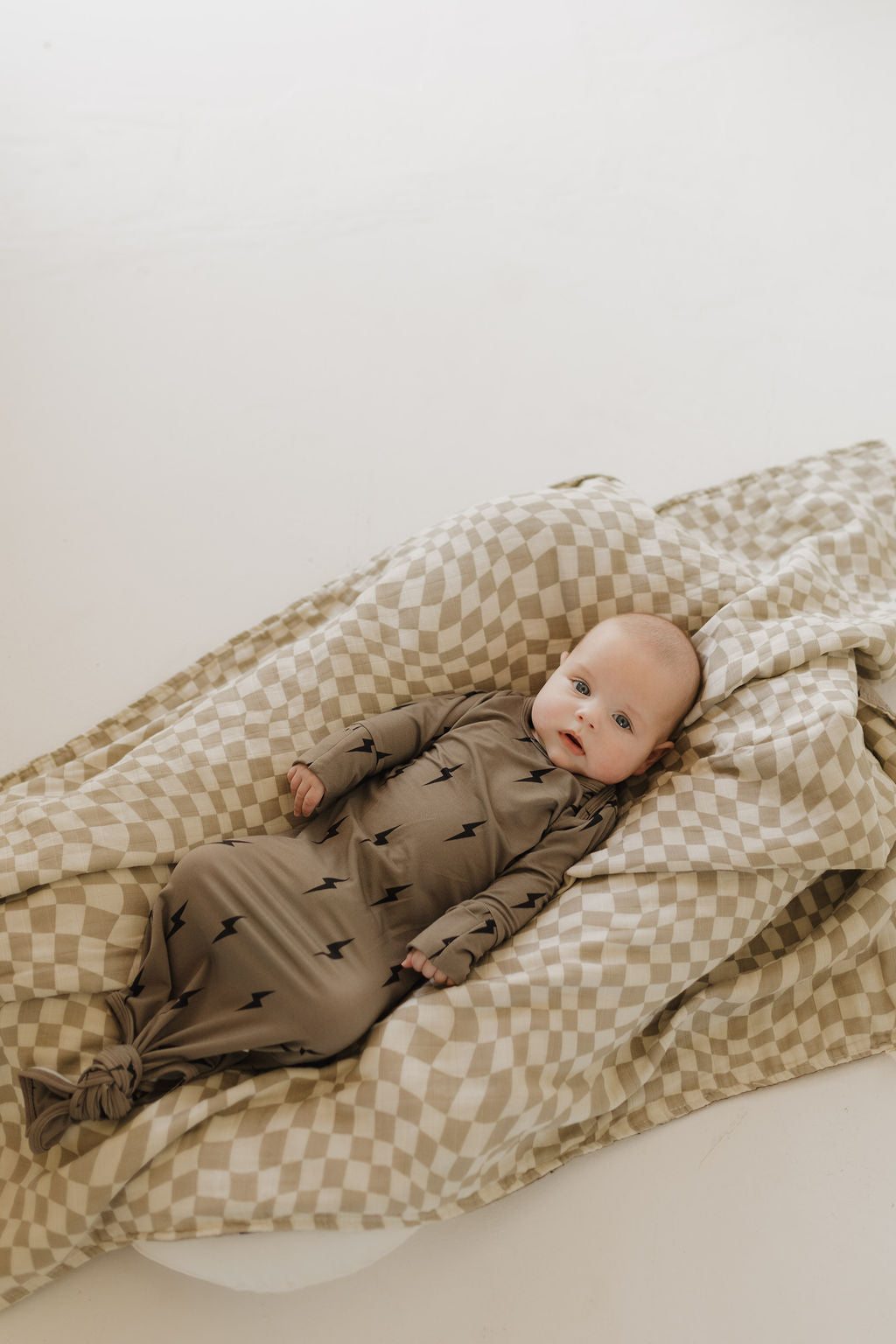 A baby lies on a soft, beige and white checkered blanket, wearing a hypoallergenic Bamboo Knotted Gown in dark brown with a minimalist lightning bolt pattern by forever french baby. The scene is serene and well-lit, creating a cozy atmosphere.