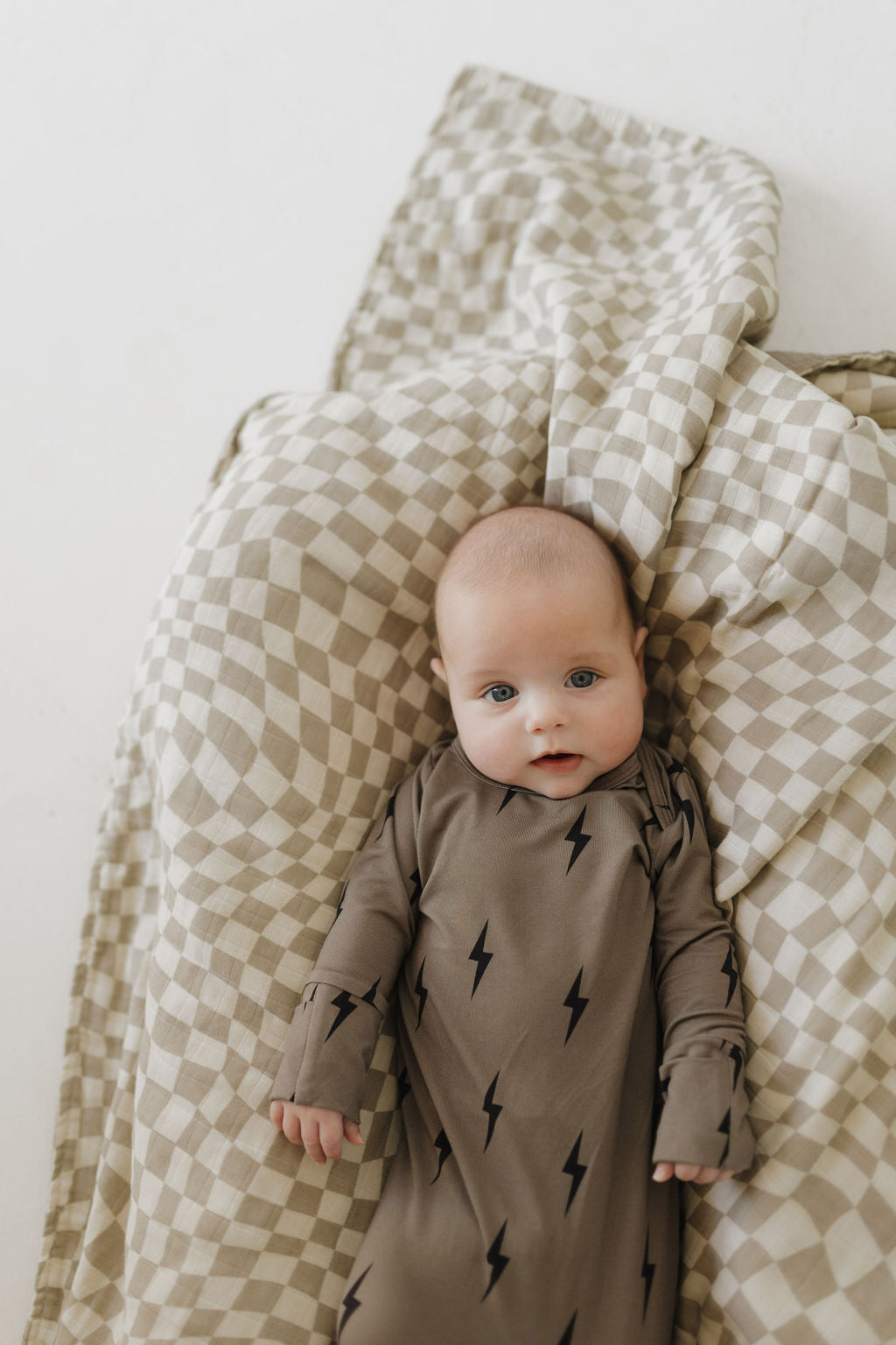 A baby in a Bamboo Knotted Gown with brown and black lightning bolts from forever french baby lies on a soft, checkered beige blanket. The breathable, hypoallergenic bamboo clothing keeps the baby comfortable as they look up with a neutral expression.