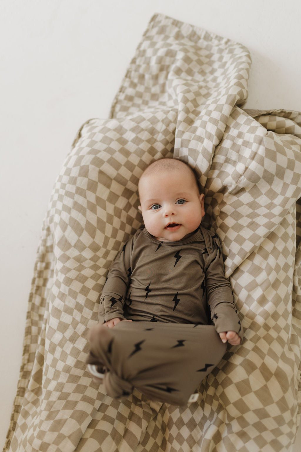 A baby in the Bamboo Knotted Gown by forever french baby, featuring a hypo-allergenic design with black lightning bolt patterns on brown fabric, lies calmly on a beige and white checkered blanket. This breathable bamboo outfit adds to the serene expression.