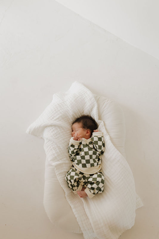 A newborn peacefully sleeps in the Sweat Set | Lucky by forever french baby, cozily wrapped in a soft white blanket on a white cushion. The light, neutral background highlights the charm of this adorable baby outfit.