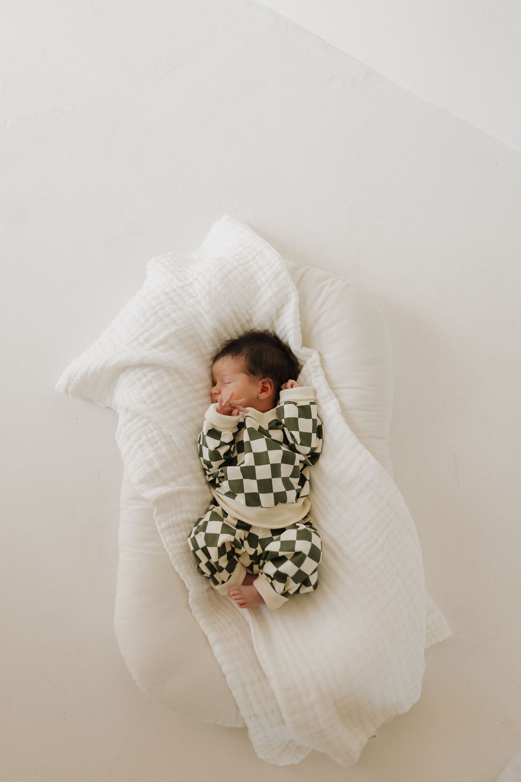 A newborn sleeps peacefully on a white cushioned blanket, wrapped in a green and white checkered romper. The baby's dark hair contrasts the neutral setting, showcasing the charm of coordinated outfits akin to the Sweat Set | Lucky by forever french baby.