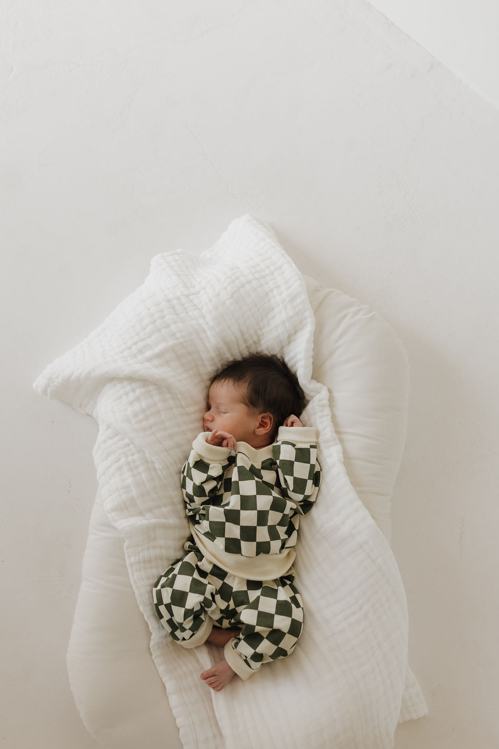 A sleeping baby in a charming green and white checkered Sweat Set | Lucky by forever french baby reclines on a soft white blanket.