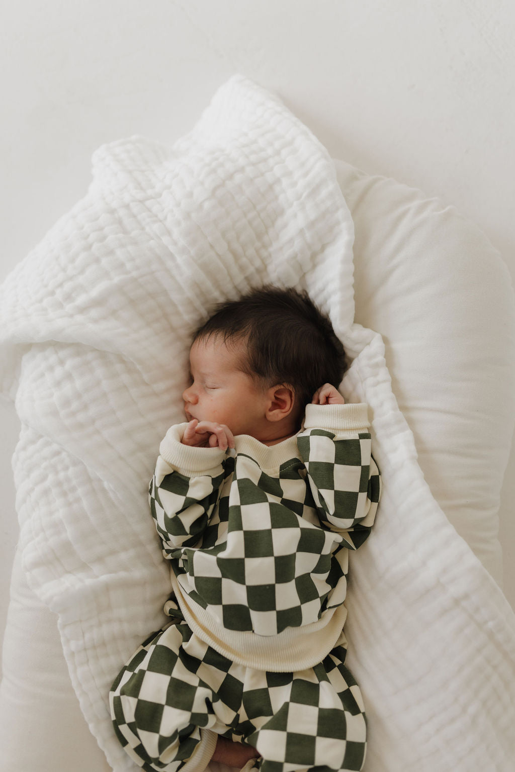 A newborn baby peacefully sleeps on a soft, white blanket in a charming green and white checkered romper. The minimalistic backdrop enhances the serene scene reminiscent of the coordinated charm found in matching sets like the Sweat Set | Lucky by forever french baby.