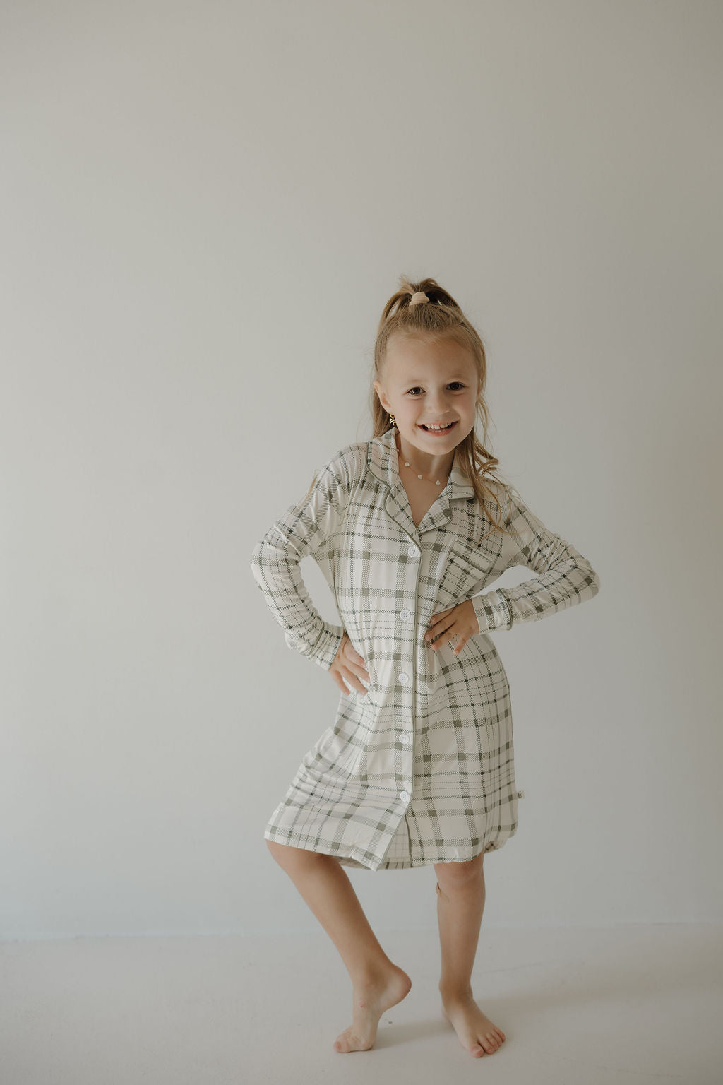 A young girl smiles barefoot with hands on hips, wearing the forever french baby Toddler Bamboo Sleeping Dress in Picnic Plaid, made from soft bamboo, against a plain light background.
