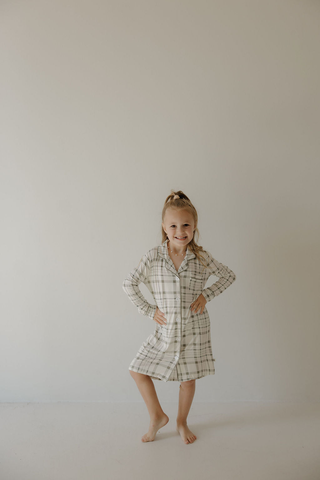 A young girl with a ponytail smiles barefoot against a plain wall, hands on hips, wearing the forever french baby Toddler Bamboo Sleeping Dress in Picnic Plaid.