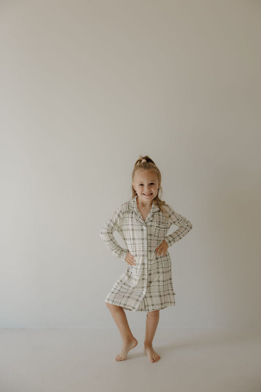 A young girl with a ponytail smiles barefoot against a plain wall, hands on hips, wearing the forever french baby Toddler Bamboo Sleeping Dress in Picnic Plaid.