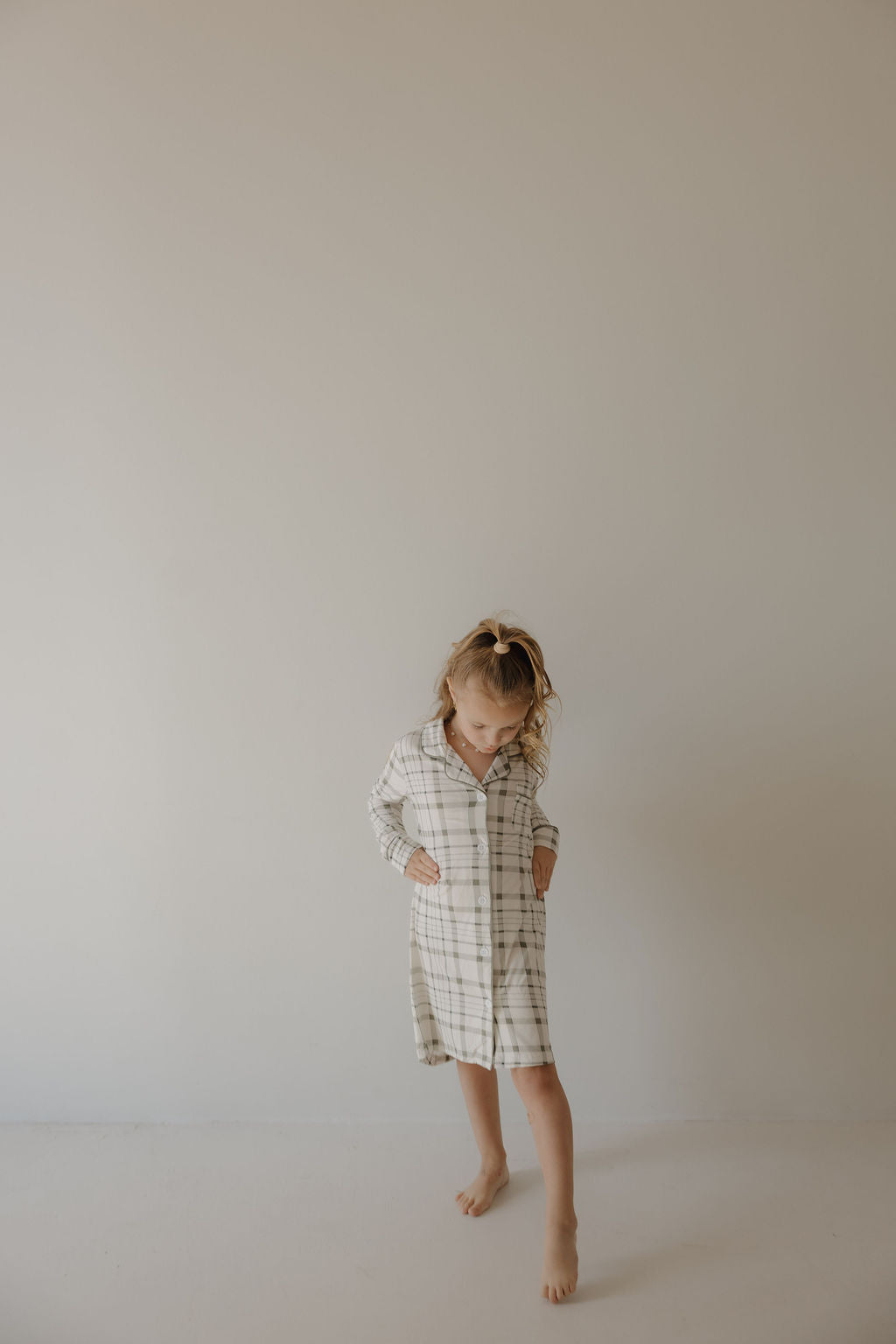 A young child stands barefoot against a plain wall, looking down and holding the sides of the forever french baby Toddler Bamboo Sleeping Dress | Picnic Plaid, a hypo-allergenic sleepwear in white and beige plaid.