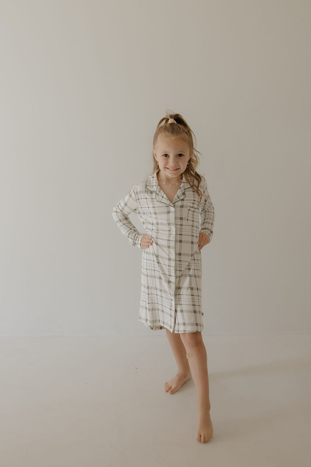 A young girl with long blonde hair in a high ponytail smiles at the camera, barefoot against a plain background, wearing the forever french baby Toddler Bamboo Sleeping Dress in Picnic Plaid.