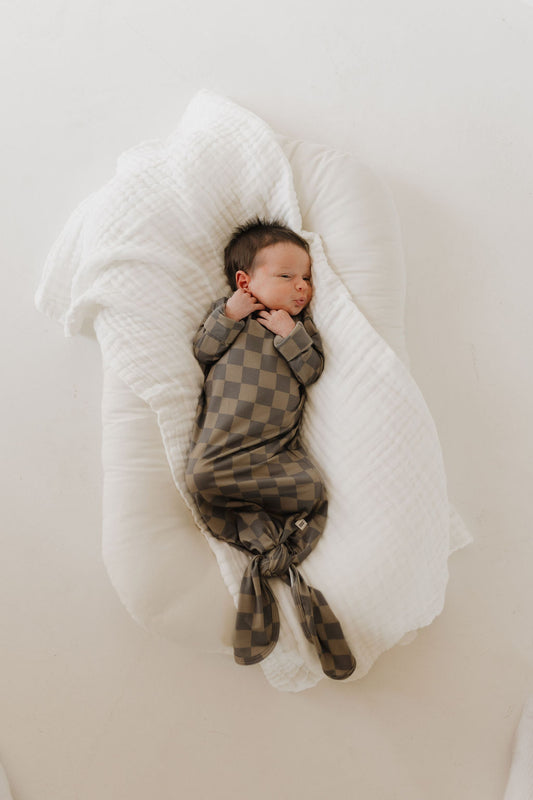 A newborn sleeps peacefully in a Bamboo Knotted Gown | Faded Checkerboard by forever french baby, wrapped in a soft white blanket on a plush cushion against a light backdrop.