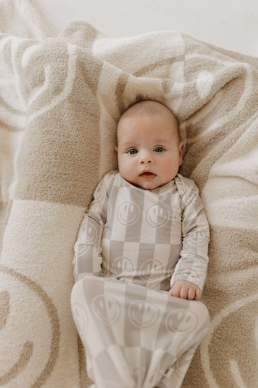 A baby lies on a beige and white patterned blanket, perfectly matched with the cozy "Bamboo Knotted Gown | Smile Checkerboard" by forever french baby. The hypoallergenic bamboo fabric and cheerful design make it ideal for delicate newborns.