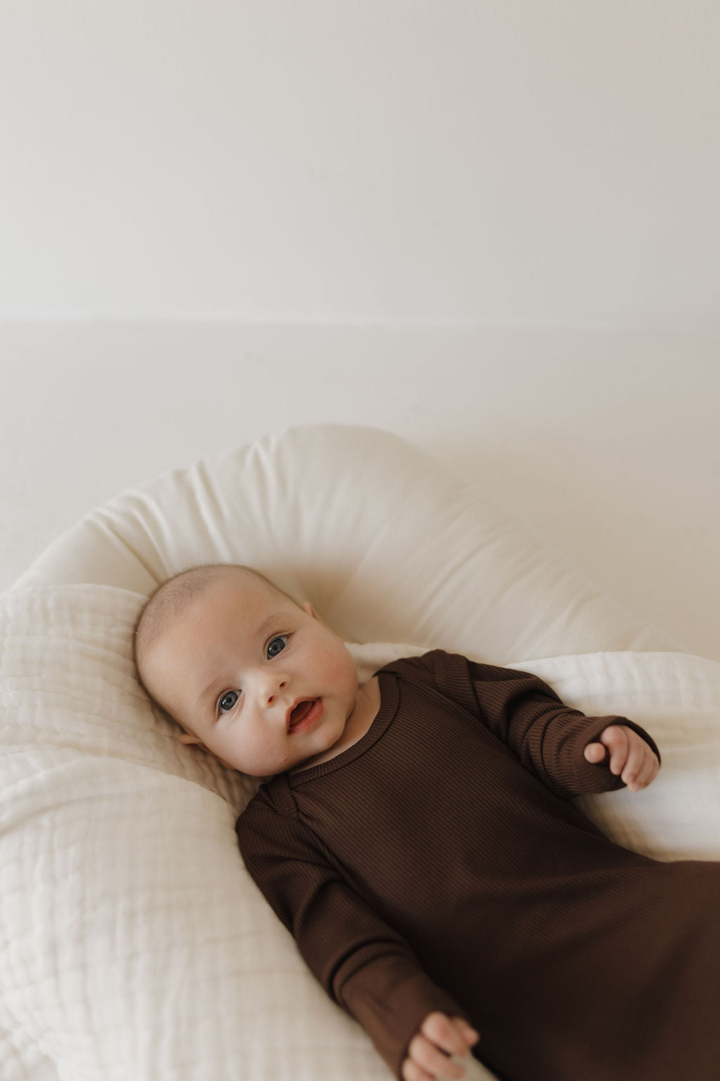 A newborn essential, the hypo-allergenic Bamboo Knotted Gown in Coffee Bean by forever french baby, cuddles the infant on a soft, white cushion. They gaze at the camera, nestled against a neutral background that radiates serenity.