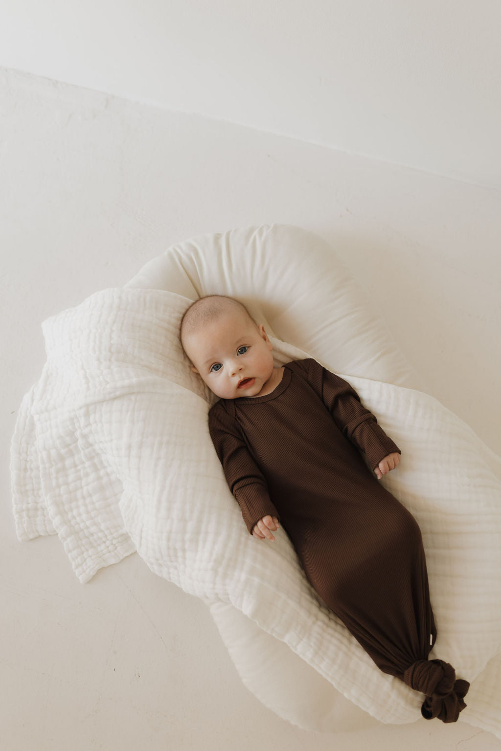 A newborn, snug in a forever french baby Bamboo Knotted Gown in Coffee Bean, lies peacefully on a hypo-allergenic cushion. The baby rests on a light-colored floor, looking directly at the camera.