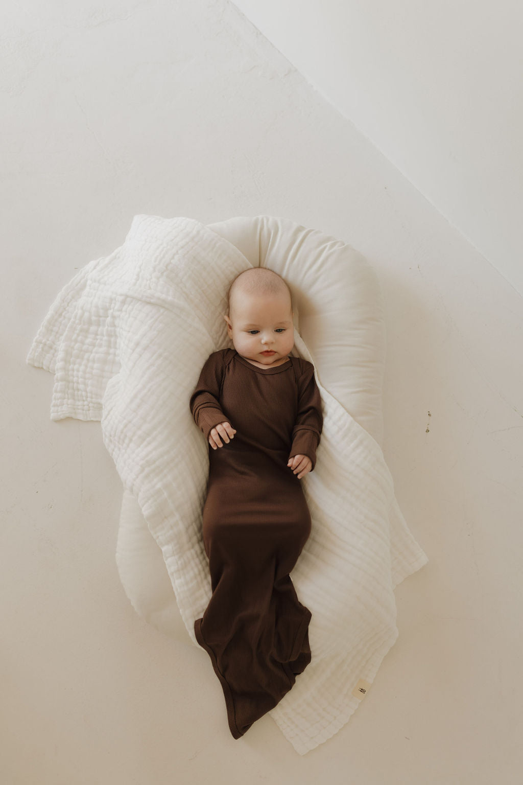 A baby in a forever french baby Bamboo Knotted Gown | Coffee Bean lies on a soft white cushion with a hypo-allergenic white blanket beneath them. The serene, minimalistic setting highlights the newborn's essential needs.