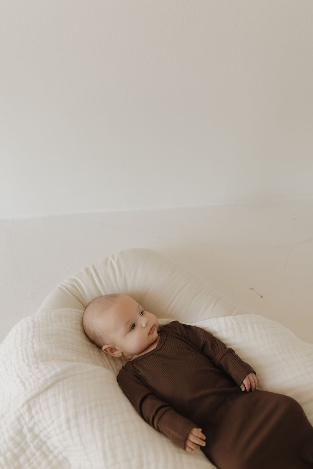 A baby wearing the Bamboo Knotted Gown in Coffee Bean by forever french baby lies on a white cushion. The hypoallergenic bamboo fabric ensures comfort and safety, with a plain, neutral background creating a serene atmosphere.