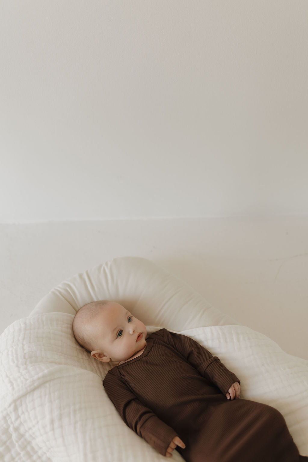 A newborn in a "Bamboo Knotted Gown | Coffee Bean" by forever french baby lies on a soft white cushion, glancing sideways. The minimalistic background and gentle lighting emphasize this essential, creating a serene atmosphere.