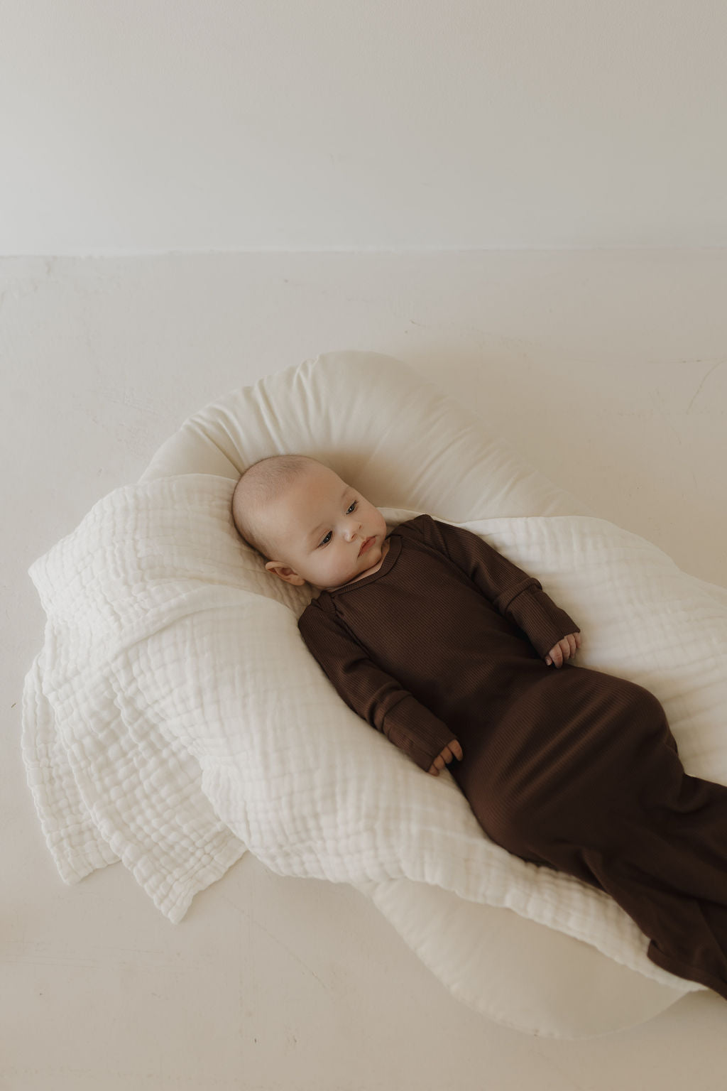 A baby in the Bamboo Knotted Gown | Coffee Bean by forever french baby rests on a cozy, white quilted blanket. The softly lit room highlights this perfect newborn essential.