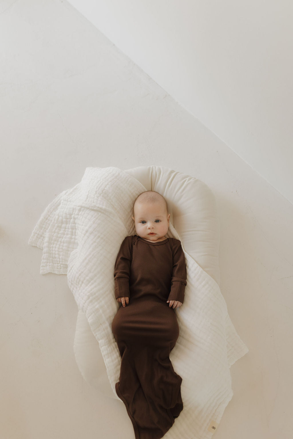 A newborn rests on a cushioned surface, wrapped snugly in the forever french baby Bamboo Knotted Gown in Coffee Bean. The hypo-allergenic fabric ensures comfort, and the light background creates a peaceful scene—a true newborn essential.