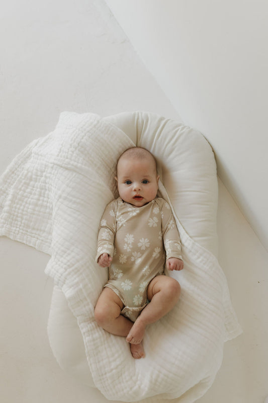 A baby wearing the "Lazy Daisy" Long Sleeve Bamboo Snapsuit by forever french baby rests on a soft, breathable cushioned mat. The backdrop is a light, neutral hue, creating a serene atmosphere.