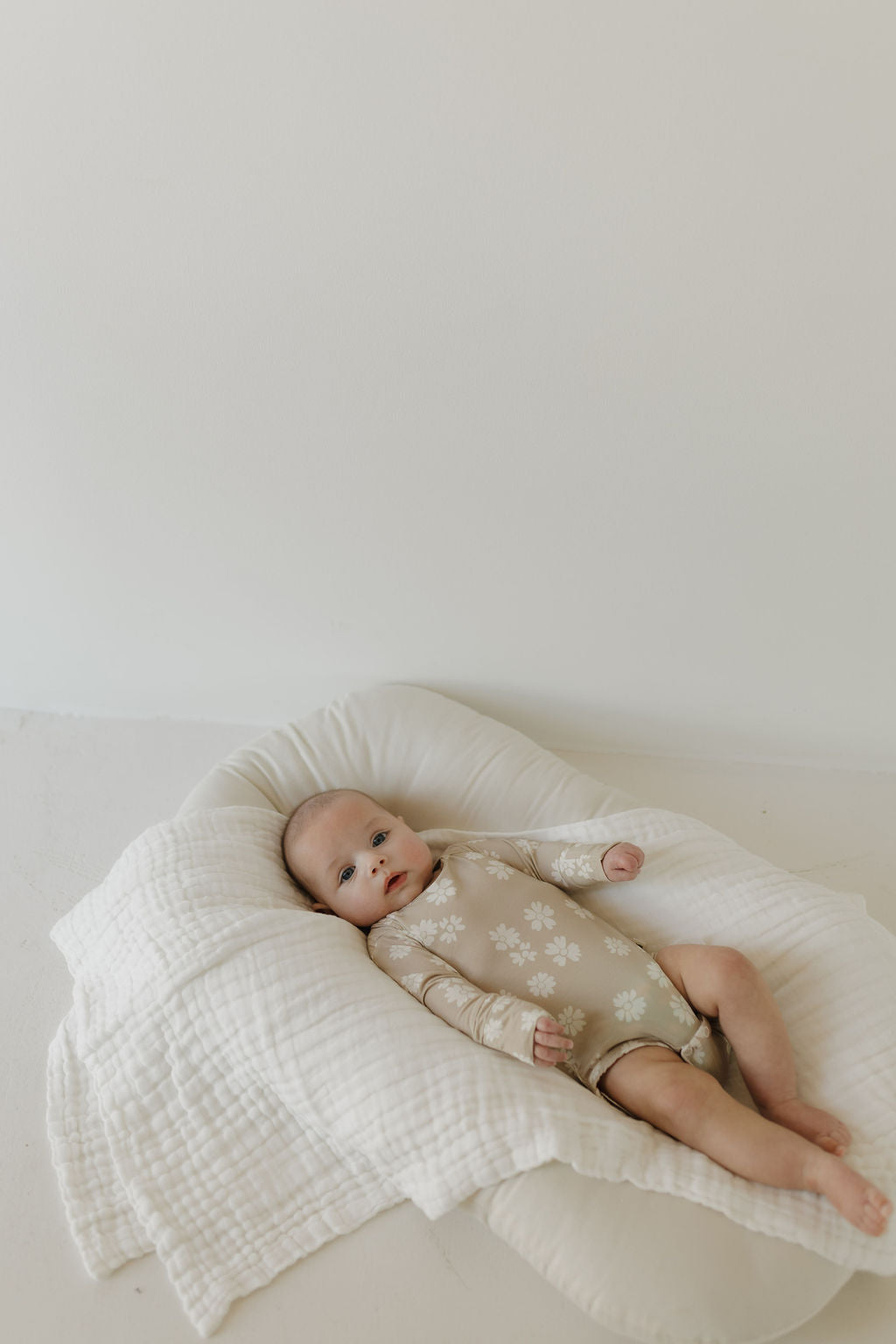 A baby wears the "Long Sleeve Bamboo Snapsuit | Lazy Daisy" by forever french baby, laying on a soft, padded white bamboo blanket. The plain white background creates a calm, serene atmosphere.