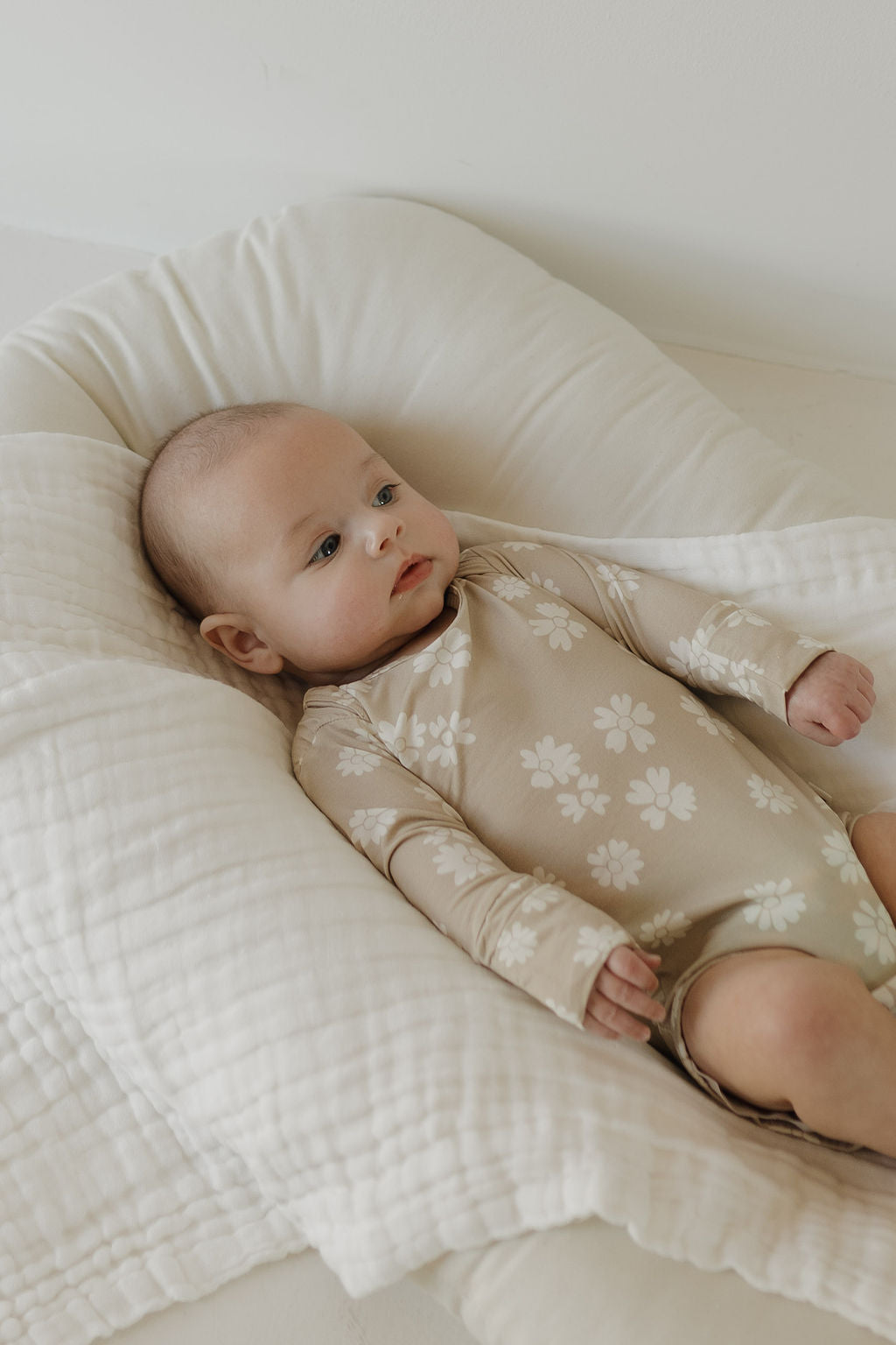 A baby in the Long Sleeve Bamboo Snapsuit | Lazy Daisy from forever french baby, featuring a beige onesie with white floral patterns crafted from breathable bamboo fabric, rests on a soft white pillow. Light reflects off the neutral background, creating a serene atmosphere.