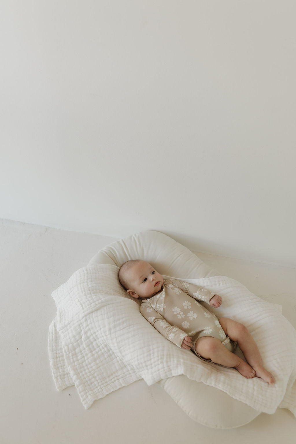 A baby in a "Lazy Daisy" beige floral bamboo snapsuit by forever french baby rests on a white cushioned blanket in a light-filled, minimalistic room, looking upwards with a calm expression.