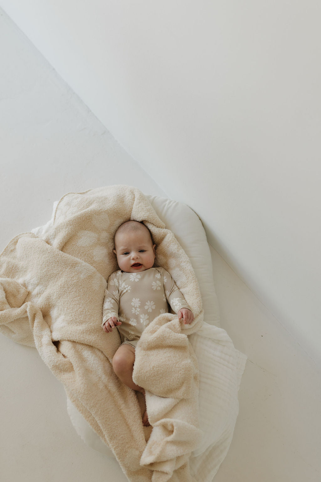 A baby is cozily wrapped in a beige, fluffy blanket on a soft, white surface. They wear the "Lazy Daisy" Long Sleeve Bamboo Snapsuit by forever french baby, featuring a light-colored floral pattern. The scene has a minimalistic pale wall background.