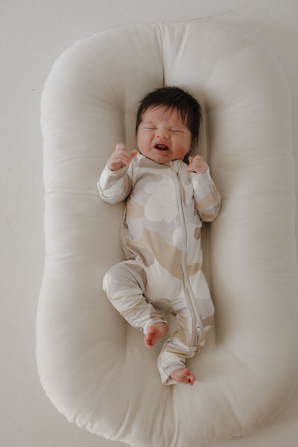A baby with dark hair lies on a soft white pad, wearing forever french baby's Bamboo Zip Pajamas | Camo 2.0, eyes closed and mouth open, appearing to cry or fuss.