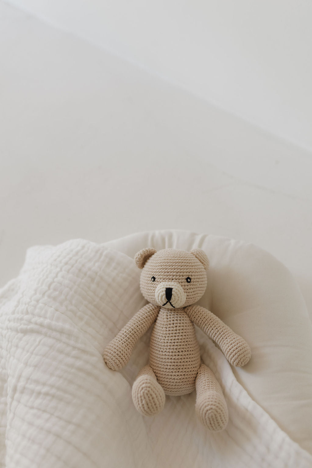 Benson the Bear, a soft knit doll from Forever French Baby, sits on a white quilt. With its light beige color and stitched facial features, this cuddly companion is positioned at the quilt's center, creating a cozy scene perfect for any nursery.