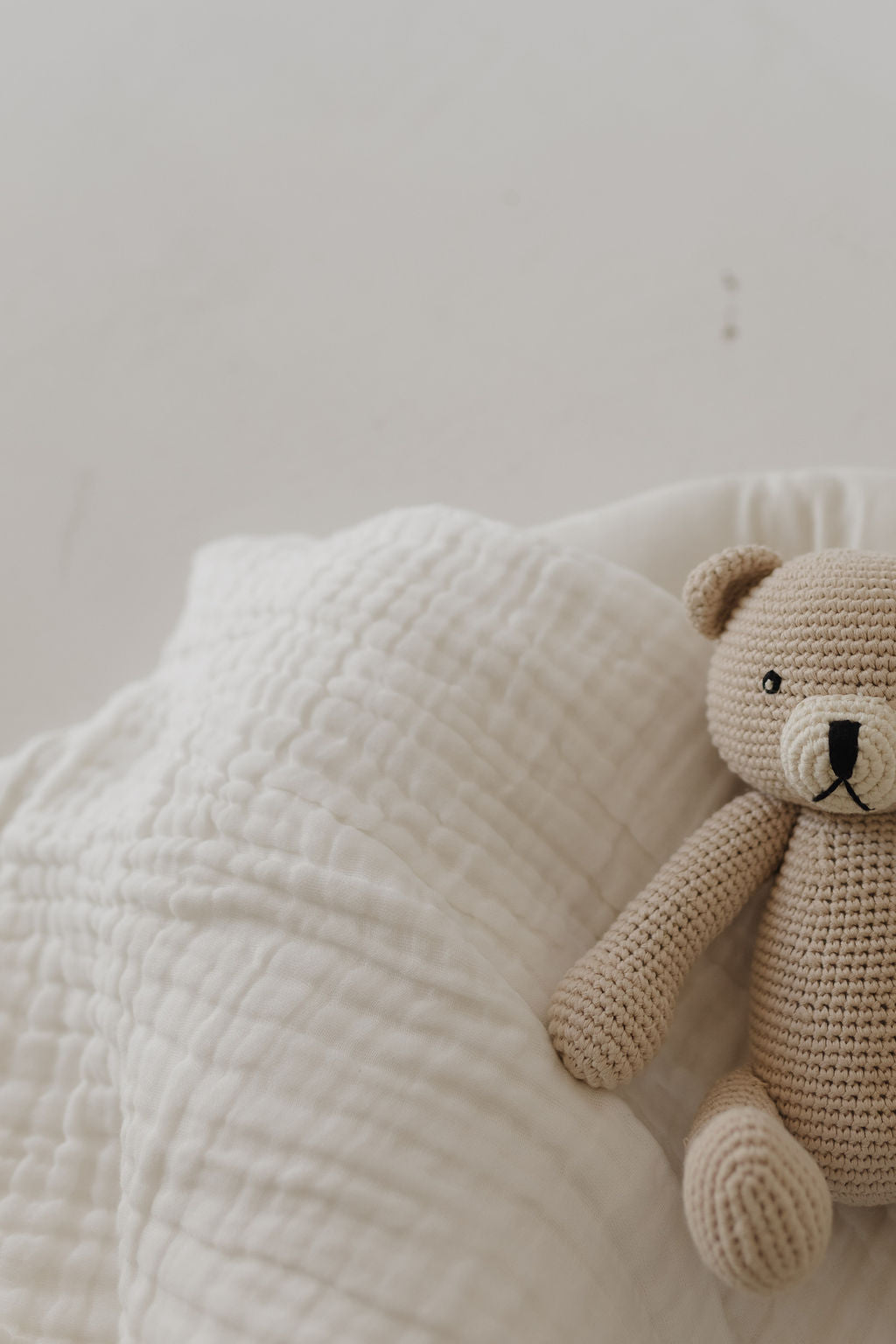 Benson the Bear, a knit doll from forever french baby, rests on a textured white blanket. Its light brown color gently contrasts against the neutral background, creating a calming and simple scene, perfect for cuddling.