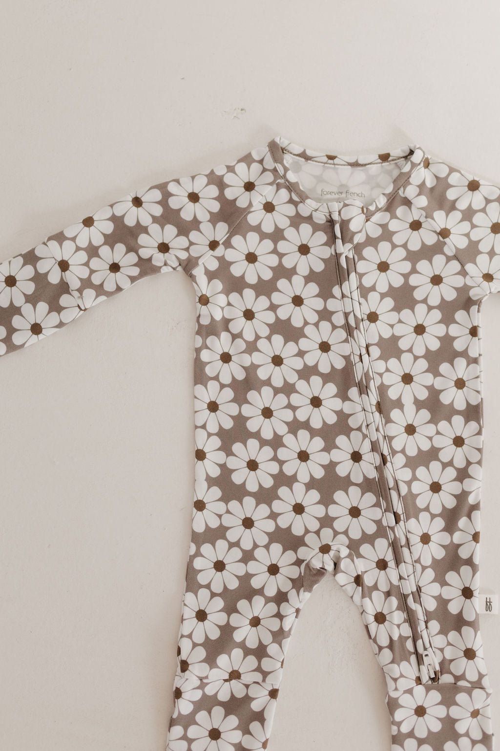 The Bamboo Zip Pajamas | Darlin' by forever french baby is a long-sleeved romper made from breathable fabric with a zipper, featuring white daisy-like flowers with brown centers on a grey-beige background.