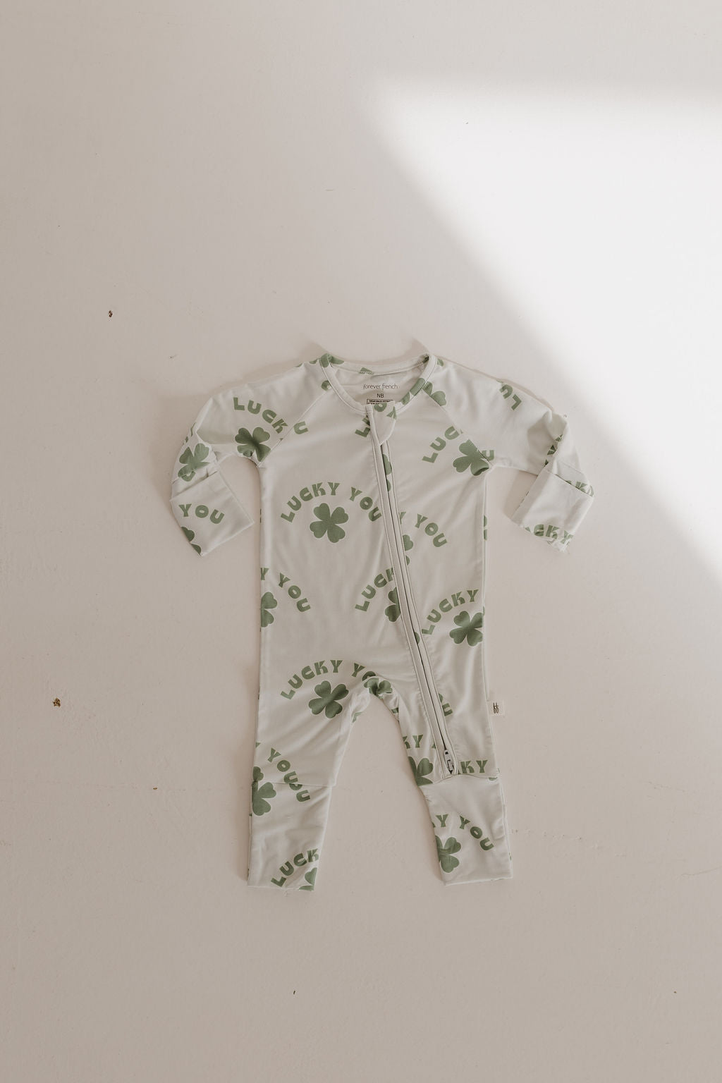 The forever french baby Bamboo Zip Pajamas in "Lucky You" feature a long-sleeve design with a front zipper, crafted from breathable, hypo-allergenic fabric. The white onesie is adorned with green four-leaf clovers and the phrase "Lucky You," displayed on a neutral background under soft lighting.