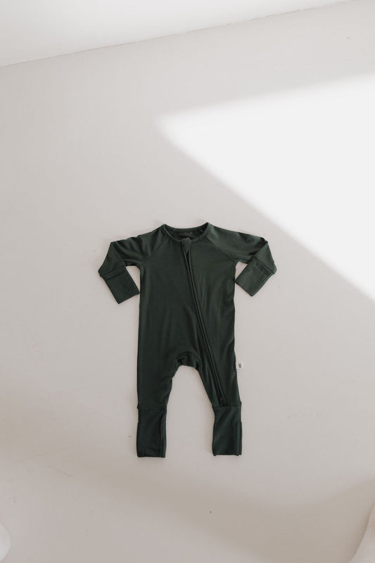 A Bamboo Zip Pajama in Emerald by forever french baby is displayed on a white surface. This hypoallergenic, breathable dark green onesie has long sleeves, legs, a front zipper with foot coverings, and soft lighting creates shadows nearby.