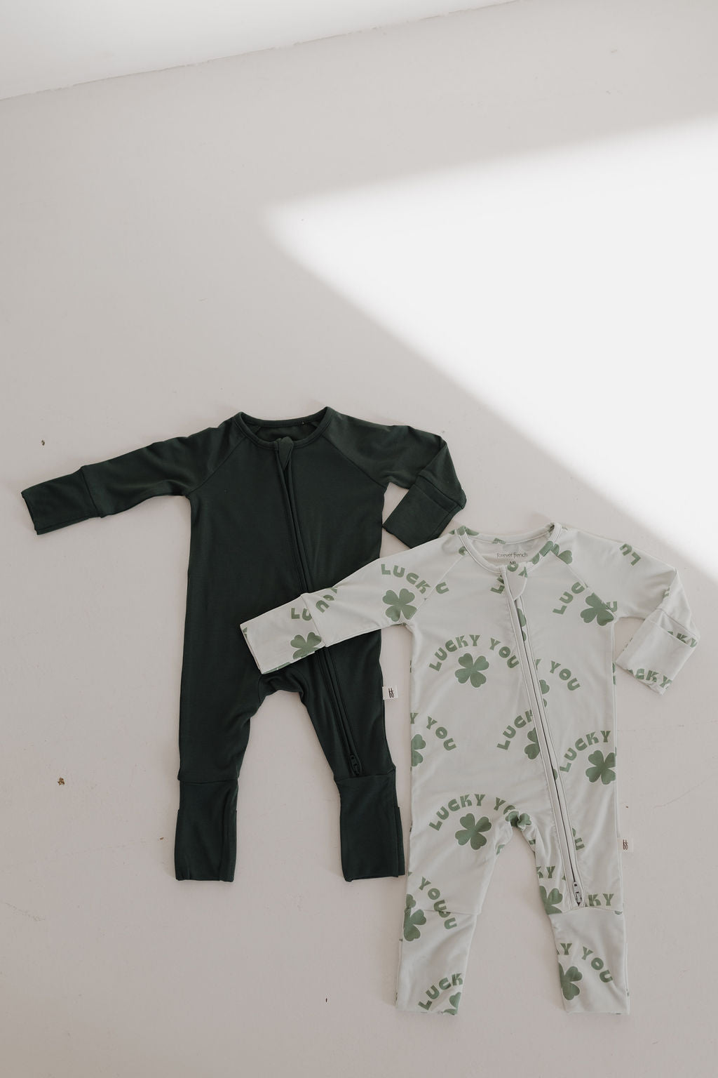 Two baby onesies rest on a light surface. One is the 'Emerald' bamboo pajama from forever french baby, featuring a front zipper in solid dark green. The other is hypo-allergenic, white with green clovers and "Lucky You" printed on it, also with a front zipper.